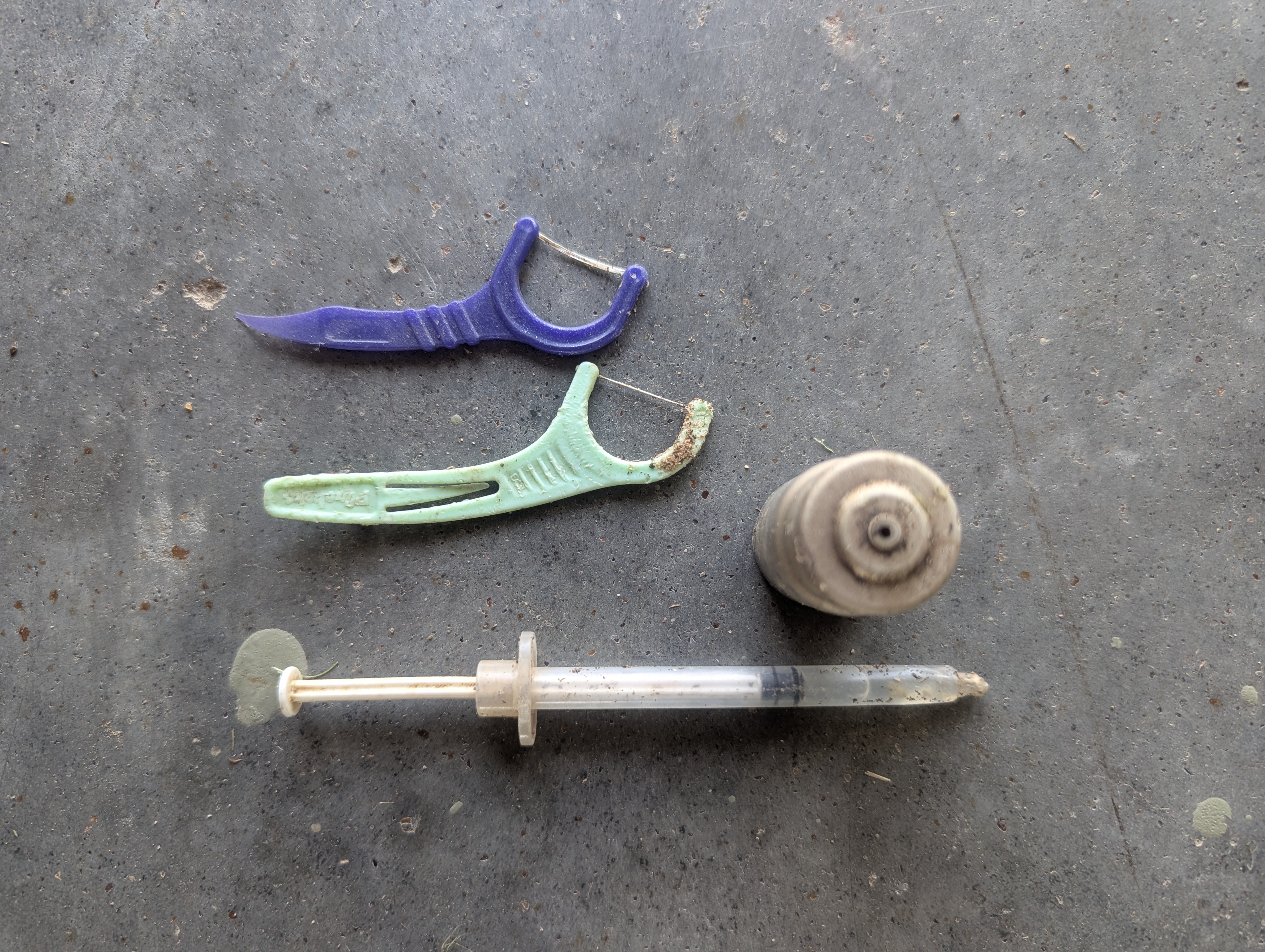 An assortment of discarded items found at a lake, including two dental flossers, a small spray canister, and a used syringe, placed on a gray surface.