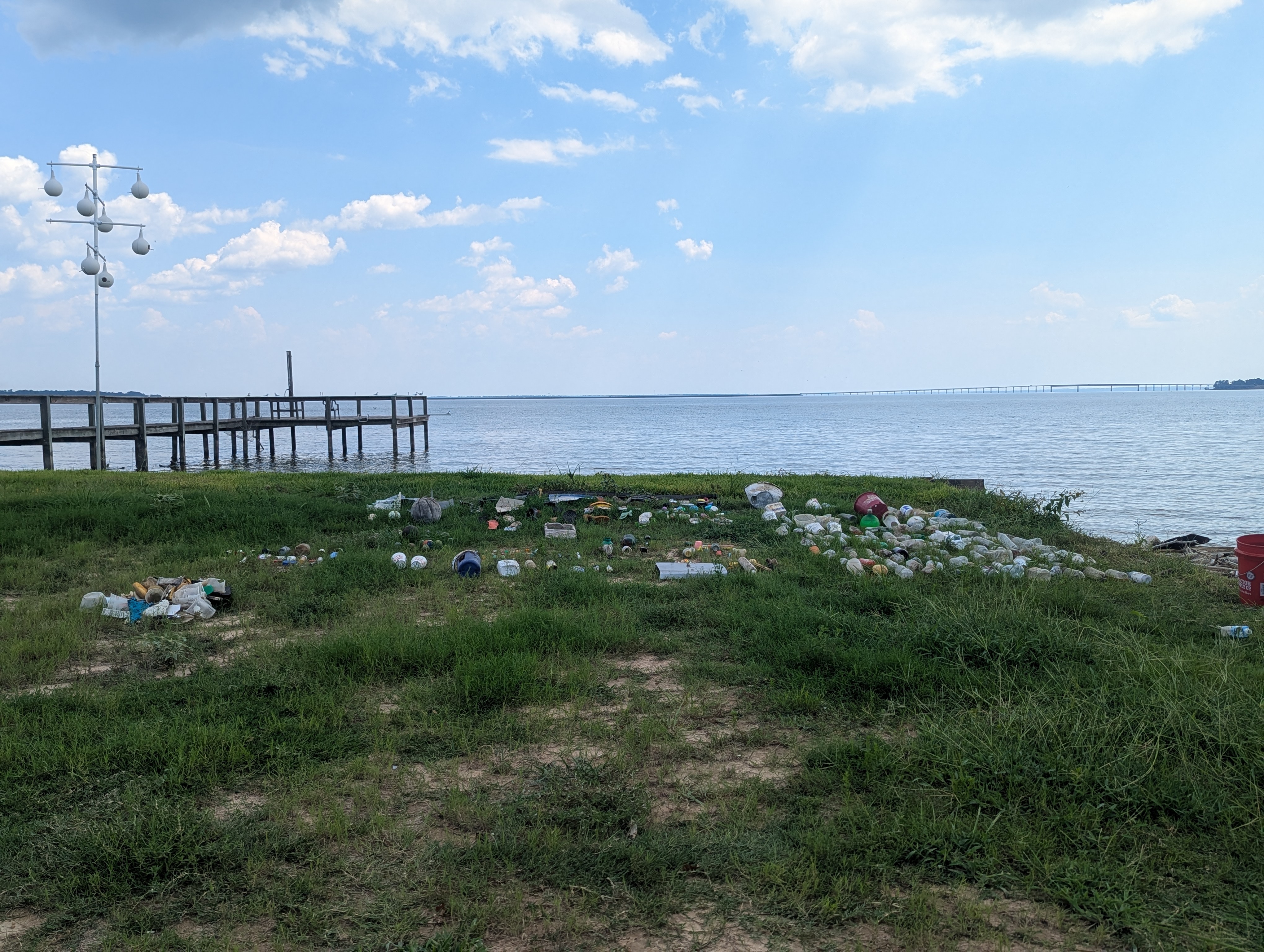 A view of a grassy area near a lake, littered with plastic bottles and other debris, alongside a wooden pier and a blue sky with clouds.