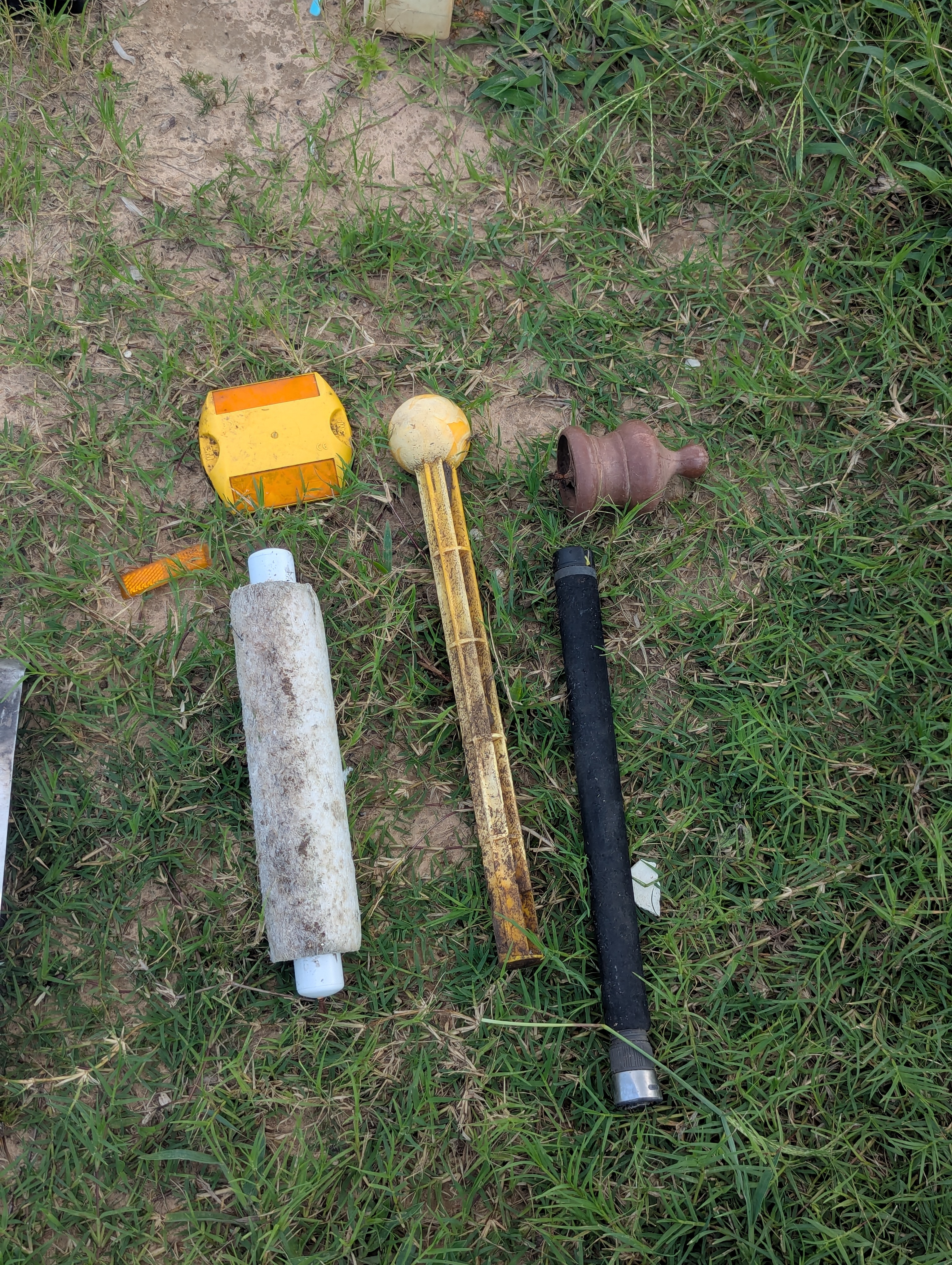 A collection of various discarded objects on grass, including a yellow object, a white cylindrical item with dirt, a yellow and brown stick-like object, and a small brown ceramic piece.